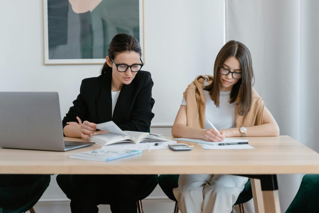 Two women working together with books and a laptop, focused on learning.