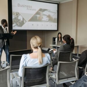 A diverse group attends a business analysis seminar with a presenter and projection screen in a modern conference room.